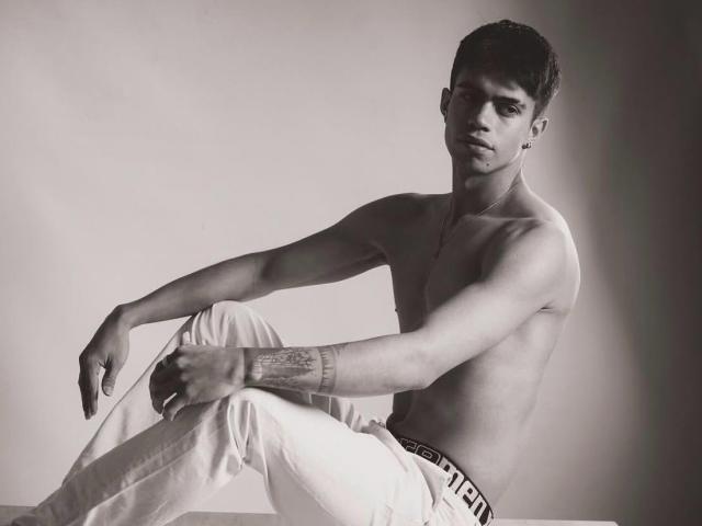 Black and white photo of CodyPipper. CodyPipper, shirtless, has a lean, muscular build with a tattoo on his right forearm. He wears white pants with a black waistband that has text. CodyPipper is seated with his left arm resting on his knee and his right arm extended forward. His short, dark hair is styled neatly. He has a serious, slightly intense expression and wears a thin necklace. The light source is to the right, creating a shadow on the left side of his body. The overall composition is simple and focused on CodyPipper's form and expression.