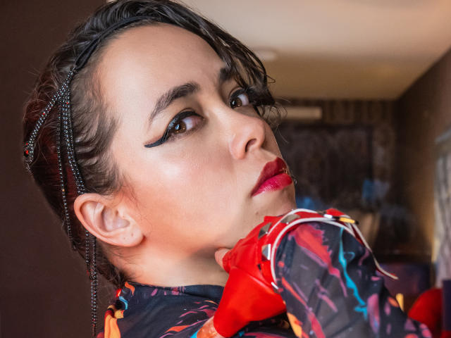 Barbara Leythonn mit kurzen, schwarzen Zöpfen, auffälligem schwarzen Eyeliner, rotem Lippenstift und einem farbenfrohen, gemusterten Oberteil, die über ihre Schulter schaut, mit roten Handschuhen. Ihr Gesicht ist leicht zur Seite gedreht..