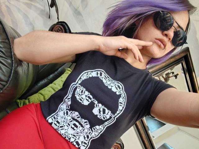 CassieJones with purple hair, black t-shirt, white skull print, red pants, black sunglasses, and finger to lips, sitting on green couch, showing off her tits.