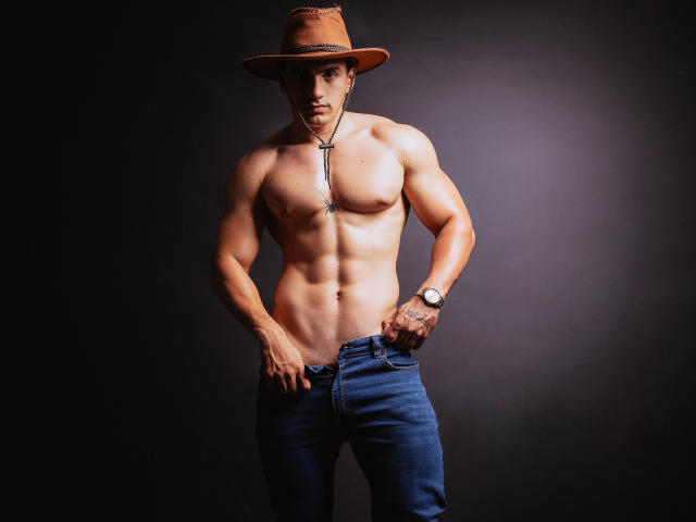 MykeOwen, shirtless with a muscular, defined, tan, and chiselled physique, wearing blue jeans, a brown hat, and a necklace, standing with hands in pockets, dark background.