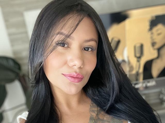 AlessandraMalory has long black hair, pink lipstick, and a tattoo on her right shoulder. She's smiling with a subtle, confident look. She's brown-skinned with dark eyes.