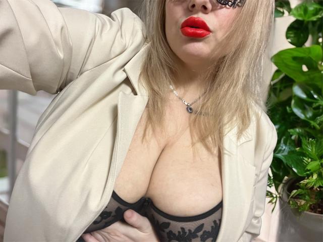 RobinLili with blonde hair, red lips, and black lace bra, showing large tits under a beige jacket, hand on her chest, green plant to the right, sexy and confident.