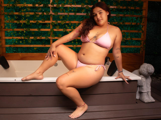 GabyCurvy with pink bikini, sitting on hot tub, one leg up, showing her ass and tits, tattoos on arm and thigh, brown hair, wooden fence, Buddha statue.