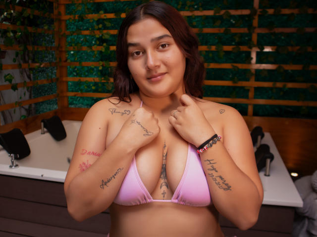 GabyCurvy with medium brown skin, dark wavy hair, pink bikini top, showing tits, tattoos on arms and chest, smiling, hands near neck, in a cozy room with a hot tub.