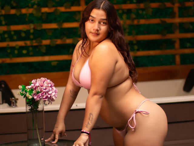 GabyCurvy with pink bikini, showing her tits and ass, leaning forward, long brown hair, tattoo on right arm, pink flowers in vase, warm indoor lighting, confident smile.