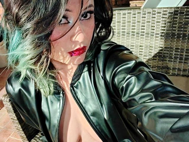 CassieJones with green and black ombre hair, wearing a black leather zip-up, exposing her tits, has red lipstick, and winks at the camera in bright sunlight.