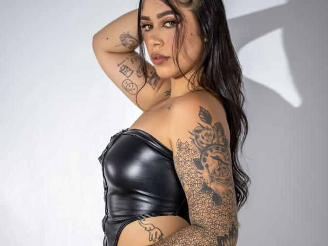CelesteSwith with tattoos, wearing a black leather strapless top, has long dark hair, brown eyes, and a serious expression. Her arm is raised, showing intricate tattoo designs.