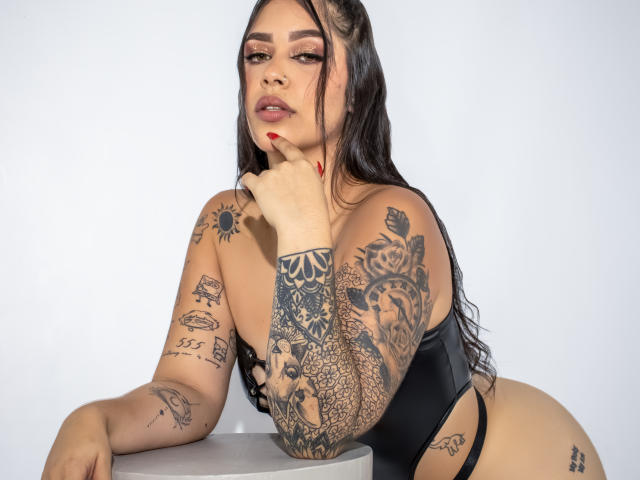 CelesteSwith with tattoos, black one-piece, leaning on table, finger on chin, red nails, long dark hair, looking at camera, sexy, full breasts, tattooed arms and legs.