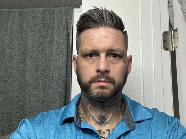 TatteDjew with short, spiked brown hair, beard, tattoos on neck and chest, wearing a blue shirt, serious expression, gray towel in background, white door with metal hinge.
