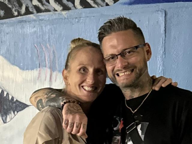 TatteDjew with blonde hair and tattoos, smiling, has arm around TatteDjew with grey hair, glasses, and beard, wearing black shirt, both smiling at camera.