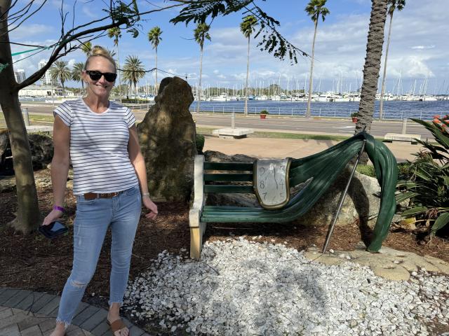 TatteDjew with blonde hair, sunglasses, striped shirt, and ripped jeans stands by a green hammock with a saddle, holding a blue phone. White pebbles and palm trees in background.
