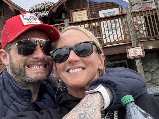 TatteDjew has tattoos, a beard, and wears a red cap with sunglasses, smiling with a blonde woman in black sunglasses and a blue jacket, holding a water bottle.