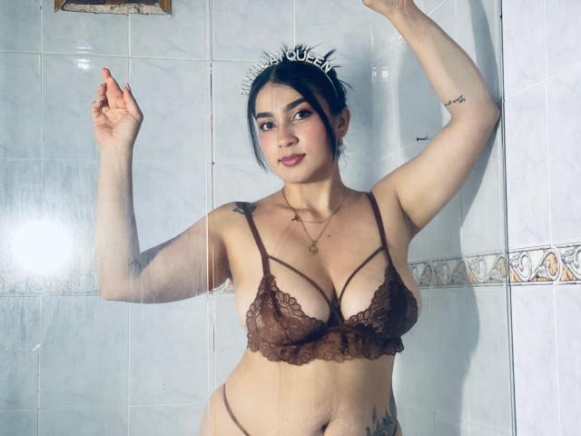 ScarletReyes with dark hair, lace brown bra, and thong, showing off her tits and a tattoo on her arm, standing against a tiled wall, right arm raised.