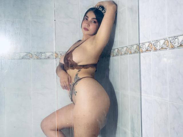 ScarletReyes with her curvy ass and big tits, wearing a brown lace bra and thong, standing in a tiled shower, one arm above her head, showing a tattoo on her side.