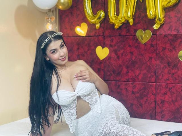 ScarletReyes with long black hair, wearing a white lace bra and skirt, touching her tits, sitting on a bed, golden balloon and heart decor in the background.