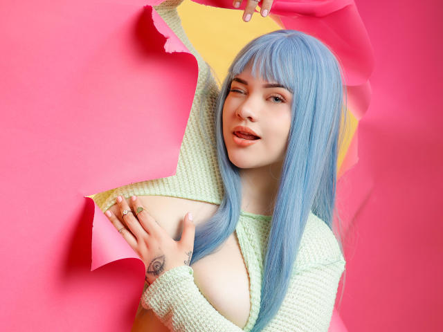 AlayaWillow, avec ses cheveux bleus, clignant de l'œil, tenant du papier rose et jaune, montrant ses seins dans un haut vert en tricot, une bague au doigt, un tatouage sur la main, fond rose..