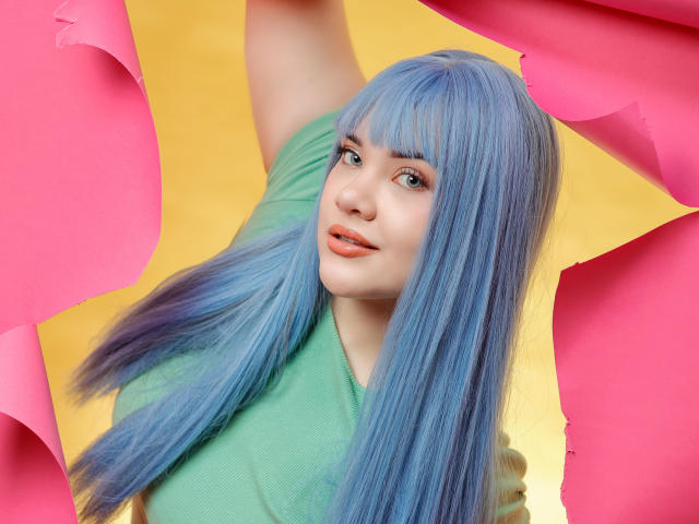 AlayaWillow with blue hair, green shirt, and orange lipstick, raising arm, surrounded by pink and yellow paper, looking at camera with a subtle smile.