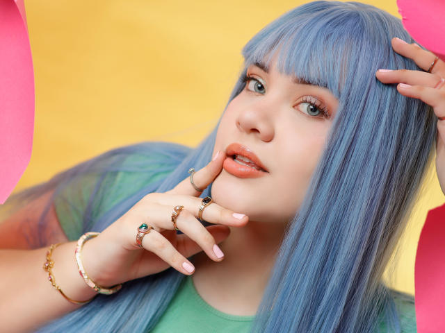 AlayaWillow with blue hair, green eyes, and pink lips, touching her chin with fingers adorned in rings and bracelets, wearing a green shirt, against a yellow background.