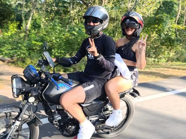 TemptationDual, un ragazzo con casco e maschera neri, maglietta nera e pantaloncini, guida una motocicletta nera con TemptationDual, una ragazza con top nero, pantaloncini bianchi e casco rosso, che fa il segno della pace..