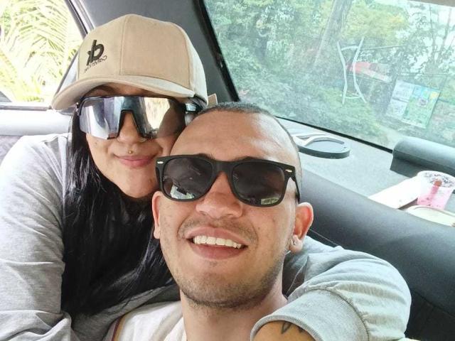 Una donna di TemptationDual, con capelli neri, piercing al naso e un berretto beige, è appoggiata a un uomo calvo e tatuato di TemptationDual che indossa occhiali da sole e sorride, seduti in macchina. Il seno della donna è premuto contro la spalla dell'uomo calvo..