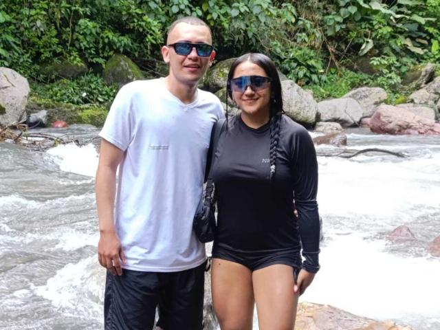TemptationDual, un ragazzo con una camicia bianca, occhiali da sole e pantaloni neri, è in piedi accanto a TemptationDual, una ragazza con una maglia a maniche lunghe nera e pantaloncini, entrambe con occhiali da sole blu, vicino a un fiume che scorre..