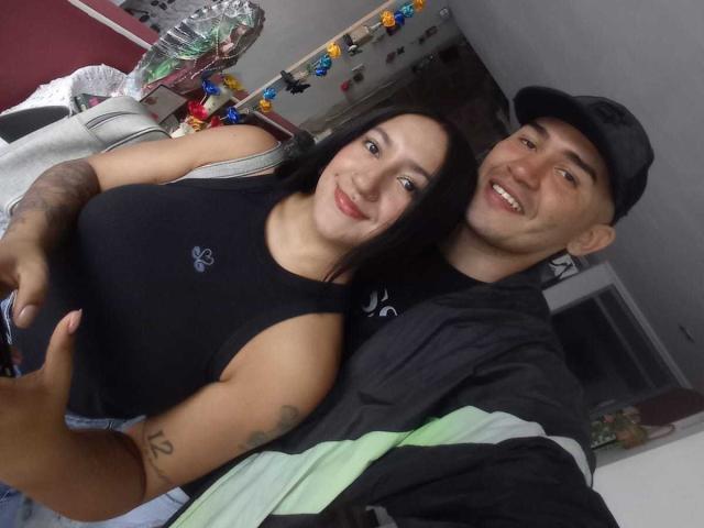 TemptationDual con il seno scoperto, indossa una canottiera nera e ha un tatuaggio, sorride, con le braccia intorno a TemptationDual che indossa una maglietta a righe verdi e un cappello nero, entrambe sorridono. Ambientazione interna, casual..