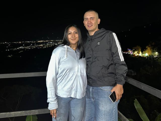 TemptationDual, con i capelli neri, seno di media grandezza e una felpa bianca, è in piedi accanto a un uomo calvo con una giacca nera Adidas, che tiene in mano un telefono. È notte, si trovano su un balcone, con le luci della città sullo sfondo..