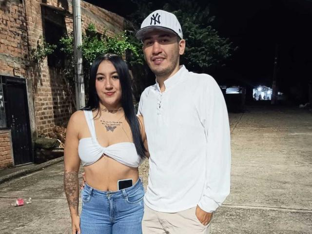 TemptationDual, con i suoi lunghi capelli neri, indossa un top bianco senza spalline che mette in mostra il seno, ha un tatuaggio sul petto e jeans blu. È in piedi accanto a un ragazzo con un berretto e una camicia bianca..