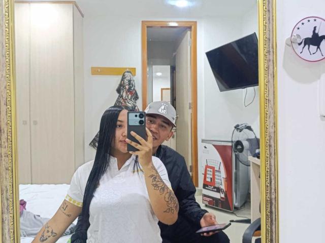 TemptationDual, con lunghi capelli neri, una camicia bianca, tatuaggi e piccoli seni, è in piedi di fronte a uno specchio, tenendo un telefono. TemptationDual, con un cappello, una maglietta nera e un sedere piccolo, è in piedi dietro..