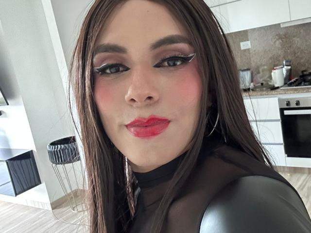 SahraValley has red lips, black eyeliner, and pink blush. She's wearing a sheer black top with long brown hair. Her tits are small, and she's smiling.