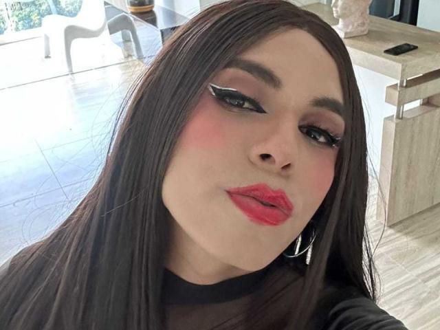 SahraValley mit langem, braunem Haar, rosigen Wangen, schwarzem Eyeliner und rotem Lippenstift, leicht schmollend, trägt ein schwarzes, durchsichtiges Oberteil und silberne Creolen..