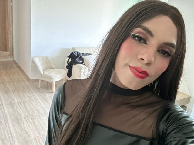 SahraValley with long brown hair, red lipstick, black sheer top, and bold eye makeup, smiling at the camera, showing her face and upper body, with a black garment in background.