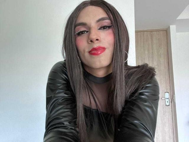 SahraValley has long brown hair, red lipstick, black leather sleeves, and pink eye makeup, with a slight smirk, wearing a black choker, in a selfie.