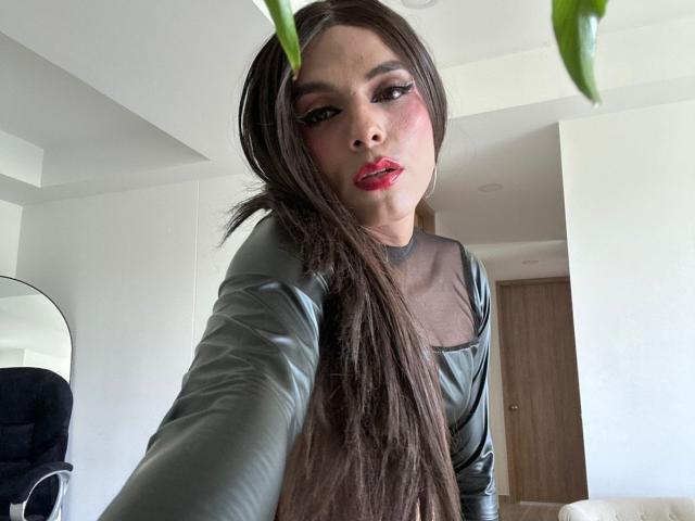 SahraValley with long brown hair, red lipstick, and green leather top, looking at camera, holding green plant leaves, in bright room.