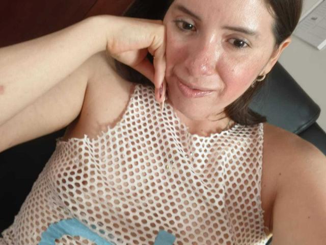 CoffeeCaress with fair skin, brown eyes, and subtle makeup, smiling at the camera. She's wearing a torn, white mesh top, exposing her small tits. Her hand rests on her cheek.