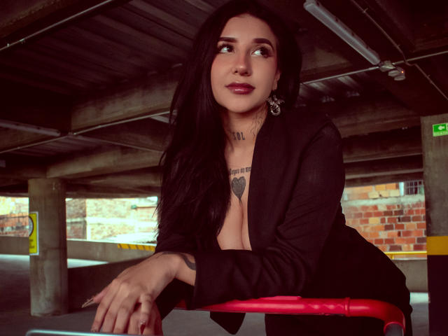 LeiaWong with long black hair, dark lipstick, and tattoos, wearing a black blazer, leaning on a red bike, under a concrete structure, looking up with a slight smile.