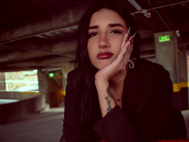 LeiaWong with black hair, red lipstick, and tattoos on her hand, resting her face on her hand in a dimly lit underground parking garage. She's wearing a black jacket.