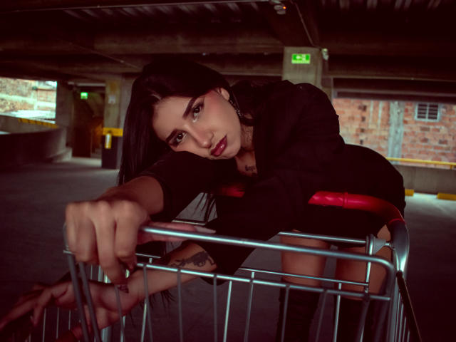 LeiaWong leans over a metal shopping cart in a dimly lit underground parking garage. Her dark hair falls over her right eye, and she gazes directly at the camera with a slightly open mouth. She wears a black top and has tattoos on her right wrist and left forearm. Her left hand grips the cart's metal frame, while her right hand rests on it. The background includes concrete pillars, an exit sign, and a brick wall. The lighting emphasizes her facial features and the texture of her hair.