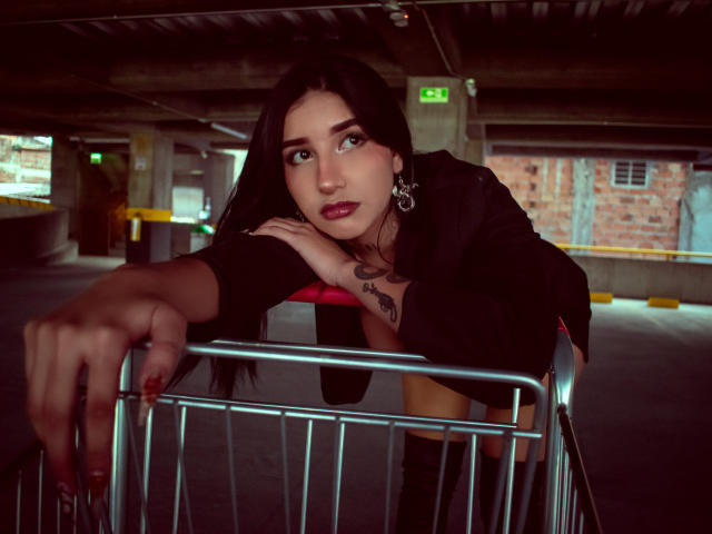 LeiaWong leans over a metal shopping cart in a dimly lit parking garage. She has long black hair, pale skin, and wears a black top with red accents, and knee-high boots. Her right hand grips the cart's top bar, while her left rests on it. She has dark, smoky eye makeup and red lipstick. A small tattoo is visible on her left wrist. She wears large, silver hoop earrings. The background shows concrete pillars and a brick wall with a yellow barrier. The image has a moody, urban atmosphere.