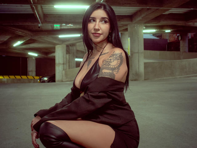 LeiaWong, squatting in a parking garage, wearing a black off-shoulder dress, showing her tits, with a dragon tattoo on her right shoulder, and black thigh-high boots.