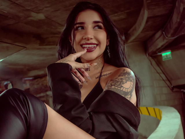 LeiaWong with long black hair, smiling, showing teeth, finger in mouth, black off-shoulder top, leather thigh-highs, tattoos, choker, looking up, under dim concrete ceiling.