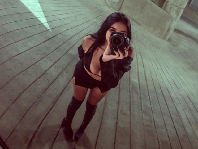 LeiaWong takes a selfie in a mirror, holding a camera in front of her face. She has long black hair and is wearing a black off-shoulder top with a deep V-neck, revealing cleavage, and black shorts. She also wears knee-high black boots. The image has a slightly greenish, muted filter. The perspective is from above, emphasizing her chest and boots. The floor is made of wooden planks.