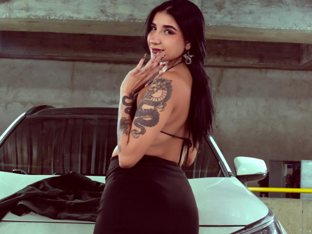 LeiaWong with black hair, backless black dress, snake tattoo on right arm, touching lips, standing in front of white car, concrete ceiling, smiling, long nails, earrings.