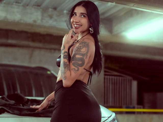LeiaWong with black hair, tattoos on her arm, smiling, wearing a black backless dress, leaning on a car, showing her ass, in a dimly lit parking garage.