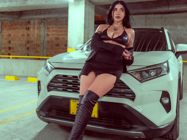 LeiaWong leans against a white Hyundai SUV in a concrete parking garage. She has long black hair, tattoos on her left arm, and wears a black bralette, short black skirt, and thigh-high black boots. Her arms are crossed, and she looks to the side with a slightly open mouth. The SUV's front grille and yellow license plate are prominent. The image is sharp with a slight sepia filter, emphasizing her bold, confident stance. The parking garage has yellow barriers and exposed brick walls in the background.