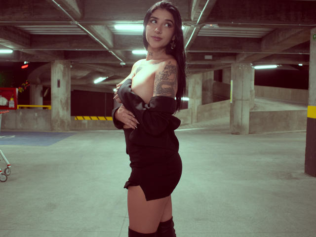 LeiaWong with long black hair, tan skin, tattoos on right arm, wearing black dress, off-shoulder, black knee-high boots, standing in dimly lit parking garage, looking to the side.