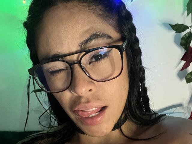 StellaBeckker with black braided hair, thick-framed glasses, and parted lips, winking, has a small nose ring, and a black choker. Her tongue is slightly out.