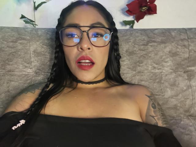 StellaBeckker with black braids, glasses, red lipstick, black choker, black strapless top, tattoo on right arm, lying on gray couch, looking at camera, slightly open mouth.