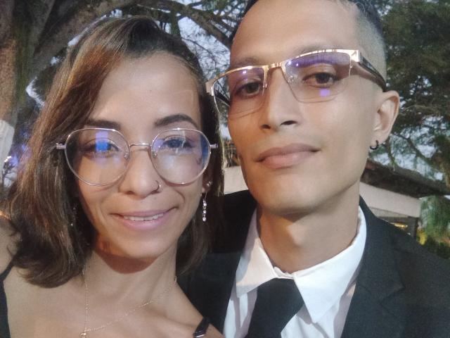 AlejoyMaria with medium tits, wearing glasses, and a black dress, next to AlejoyMaria with a bald head, glasses, and a black suit, smiling outdoors.