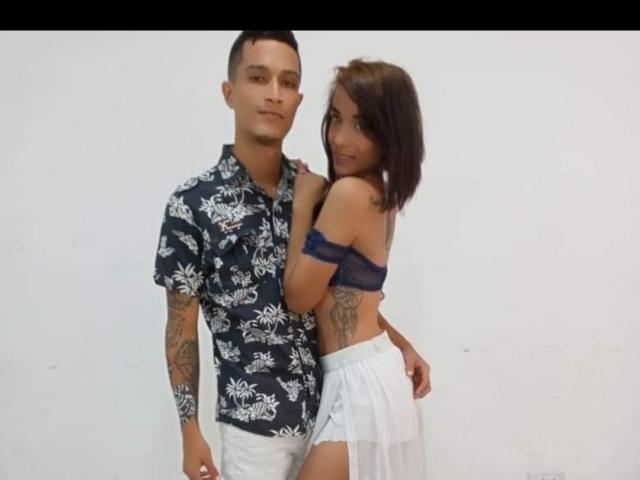 AlejoyMaria with short black hair, tattoos, wearing a floral shirt and white pants, stands with his arm around AlejoyMaria, who has brown hair, small tits, and a blue off-shoulder top.